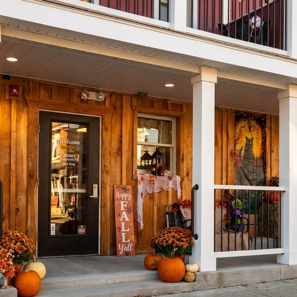 A wooden building with a porch decorated for fall with pumpkins, mums, and a "Happy Fall Y'all" sign.