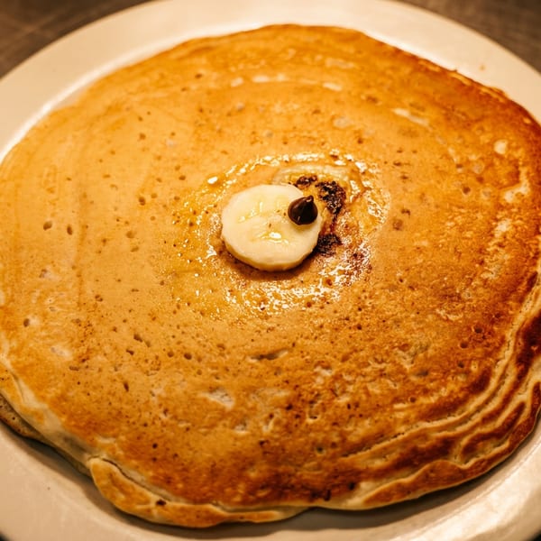 Golden-brown pancakes topped with a banana slice, chocolate chip, and syrup on a white plate.