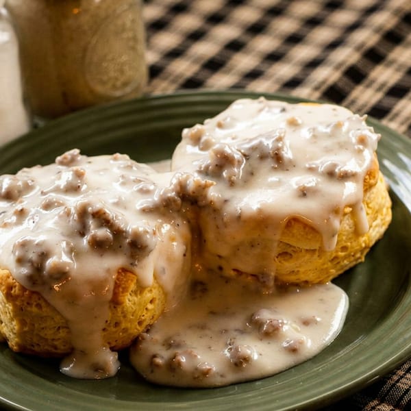Two golden biscuits covered in creamy sausage gravy on a green plate with a fork.