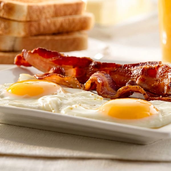 A breakfast plate with two fried eggs, crispy bacon, toast, and a glass of orange juice.